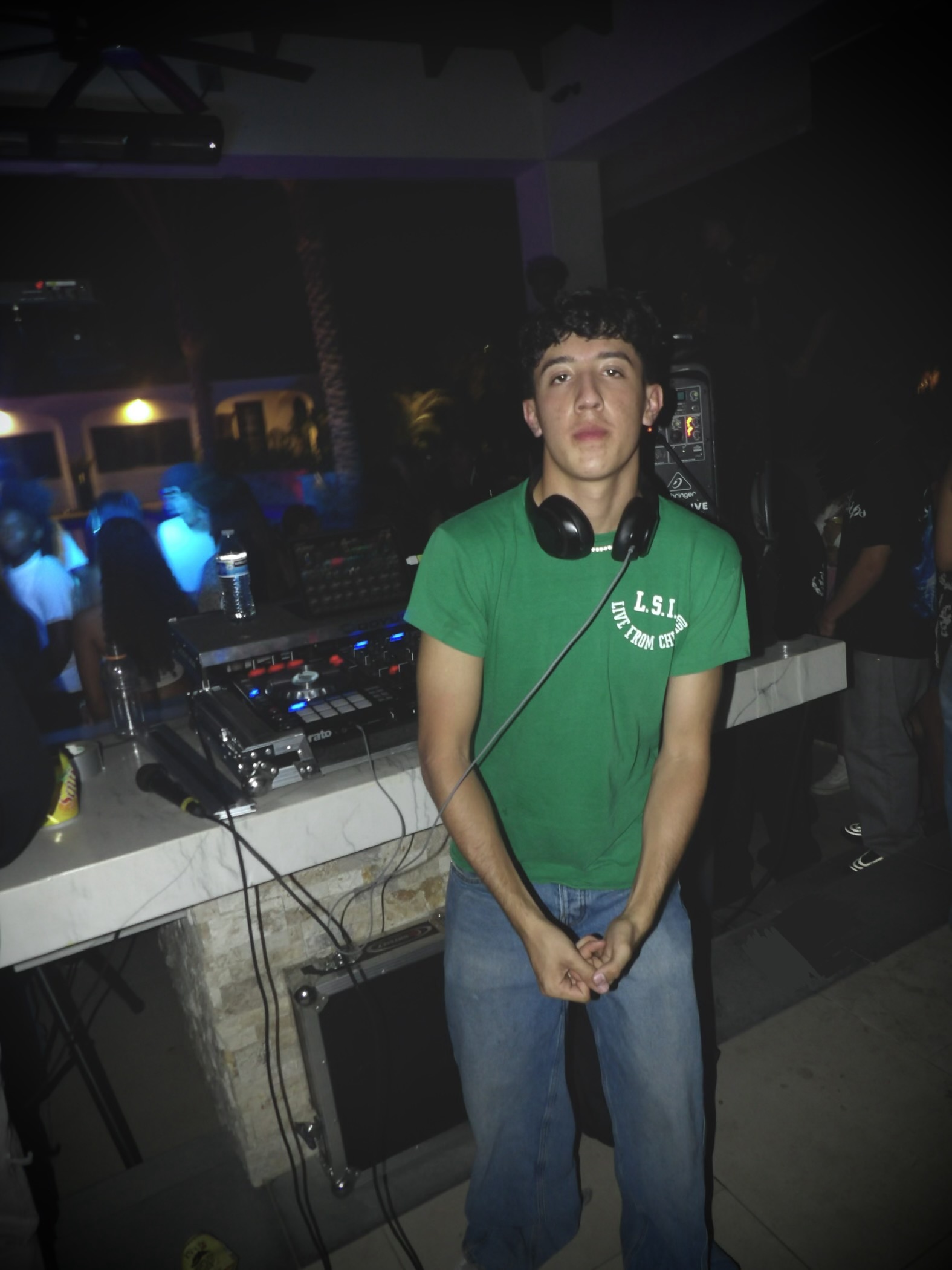 DJ Balcázar standing beside his DJ setup at a nighttime party, headphones around his neck.