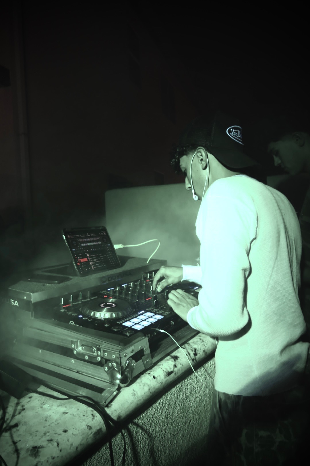 DJ Balcázar mixing on his controller in a fog-lit room, headphones to one ear.